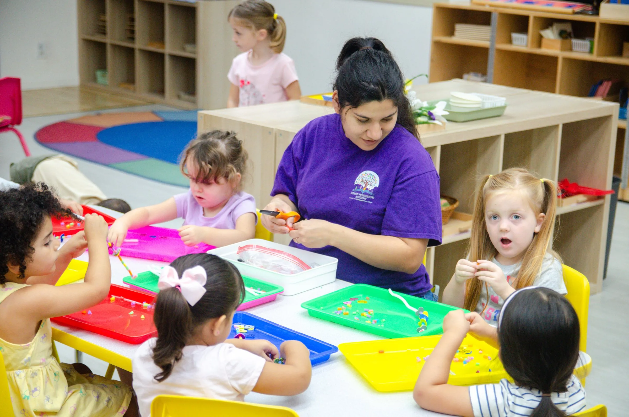 A teacher in a purple school shirt works with children at a table of colorful trays while a girl looks on in surprise.