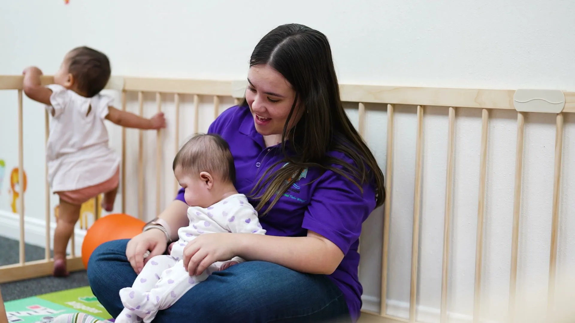 A teacher in the infant room smiles as she holds a baby on her lap while a toddler stands at the rail beside them.