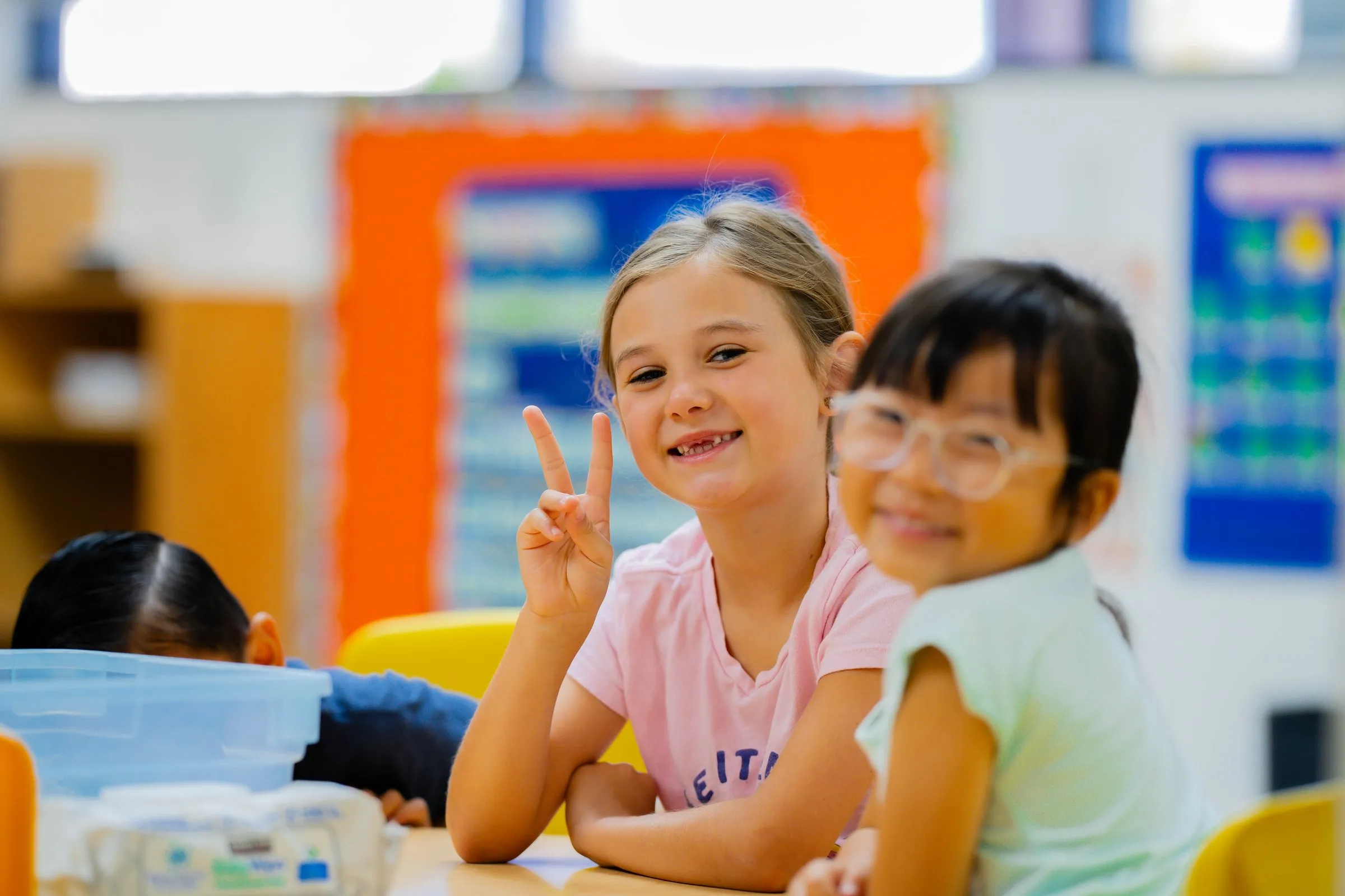 Two smiling girls sit at a classroom table, one making a peace sign.