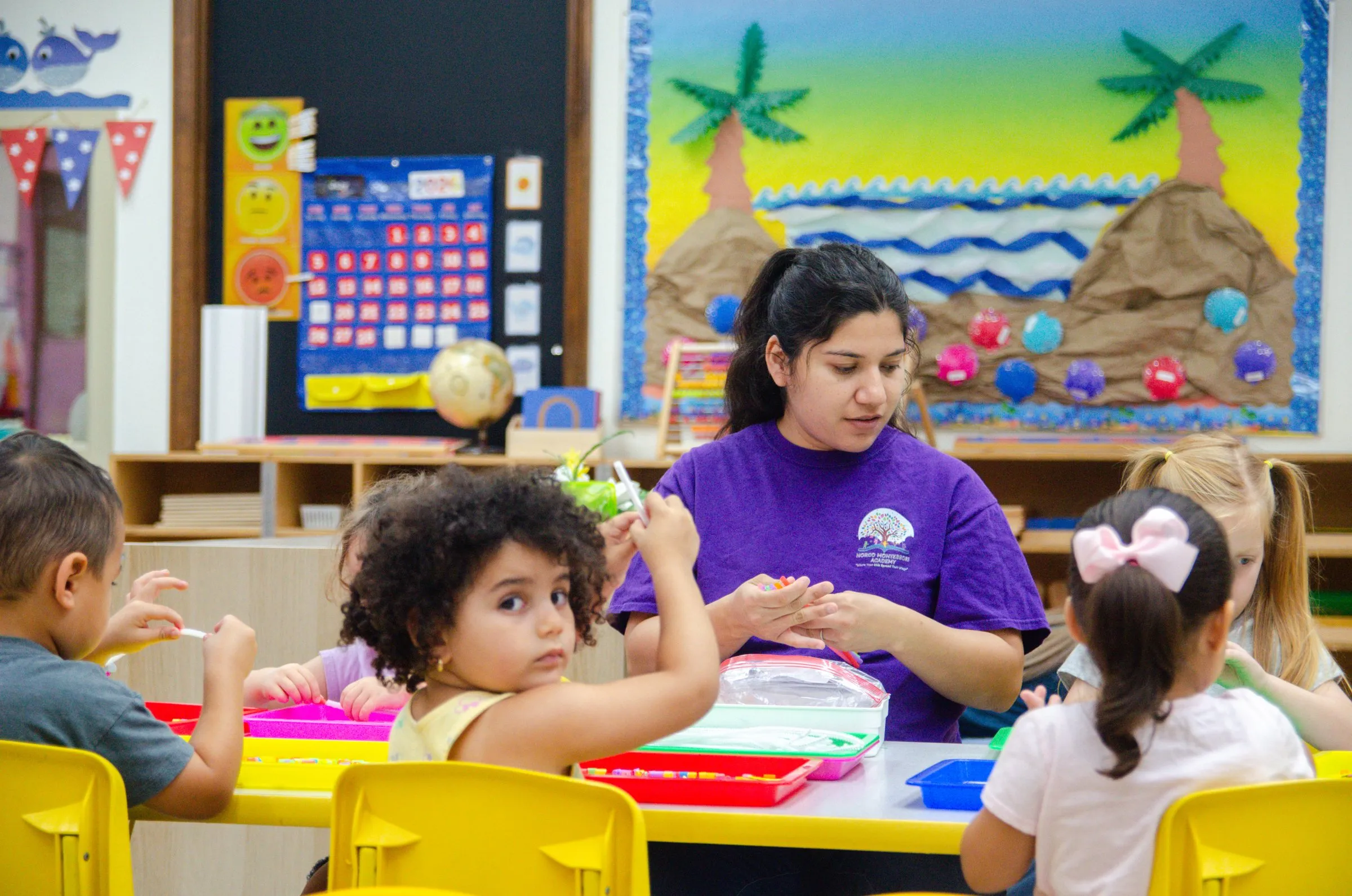 A teacher in a purple school shirt works with children at a table covered in colorful trays.