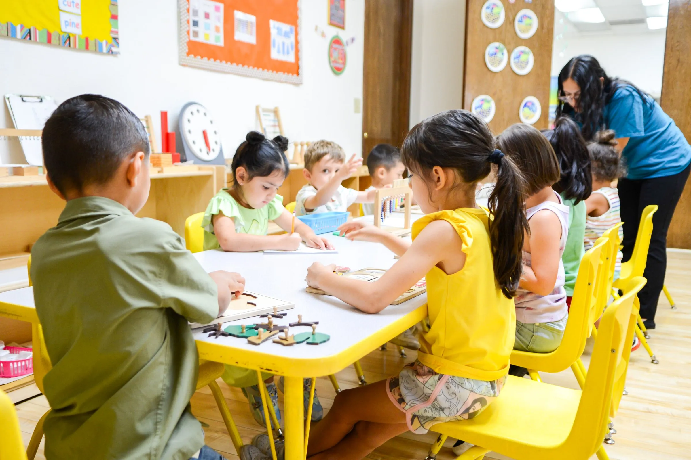 Children work at their tables with wooden puzzles while a teacher helps nearby.