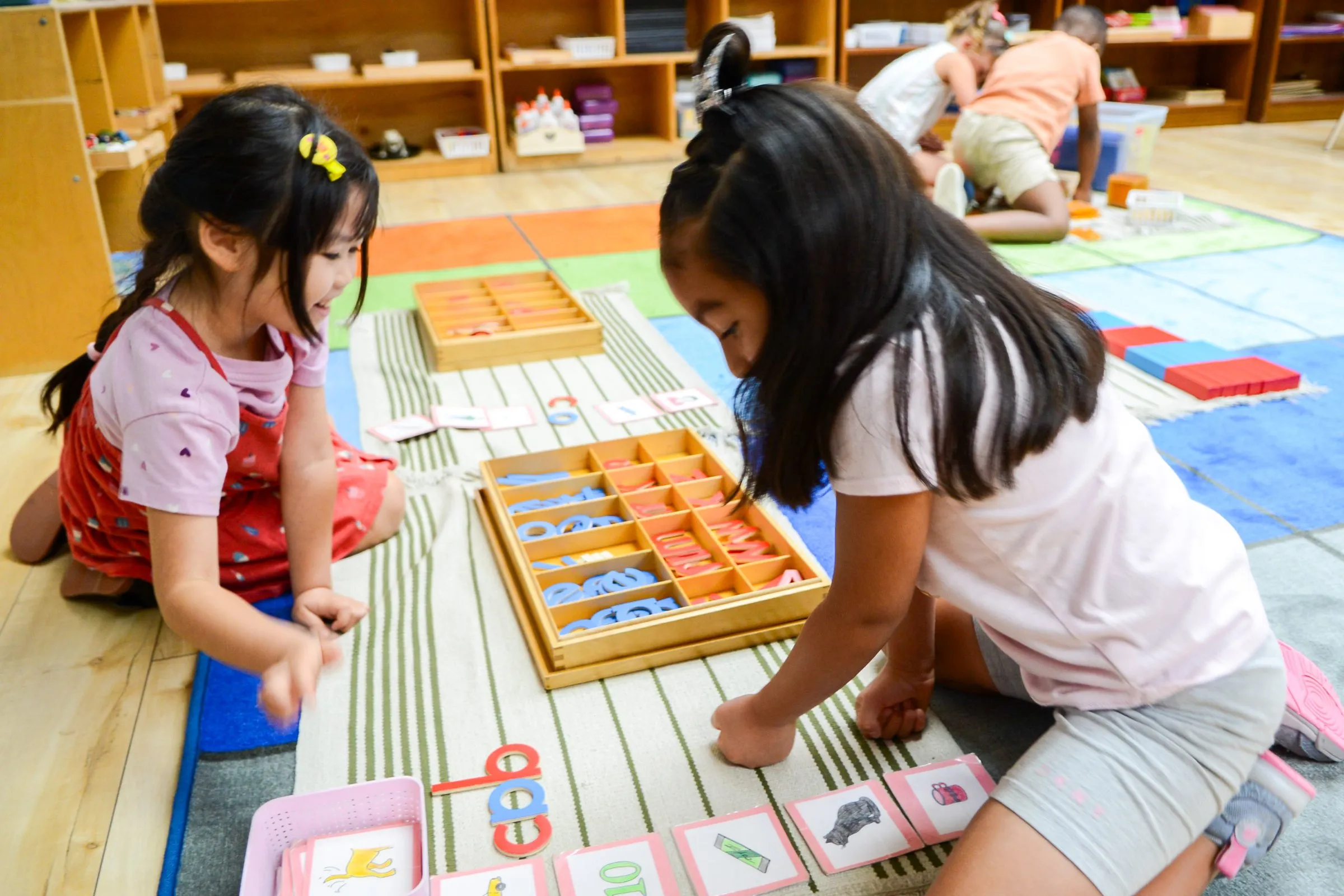 Two girls kneel on the classroom floor building words with a Montessori movable alphabet.