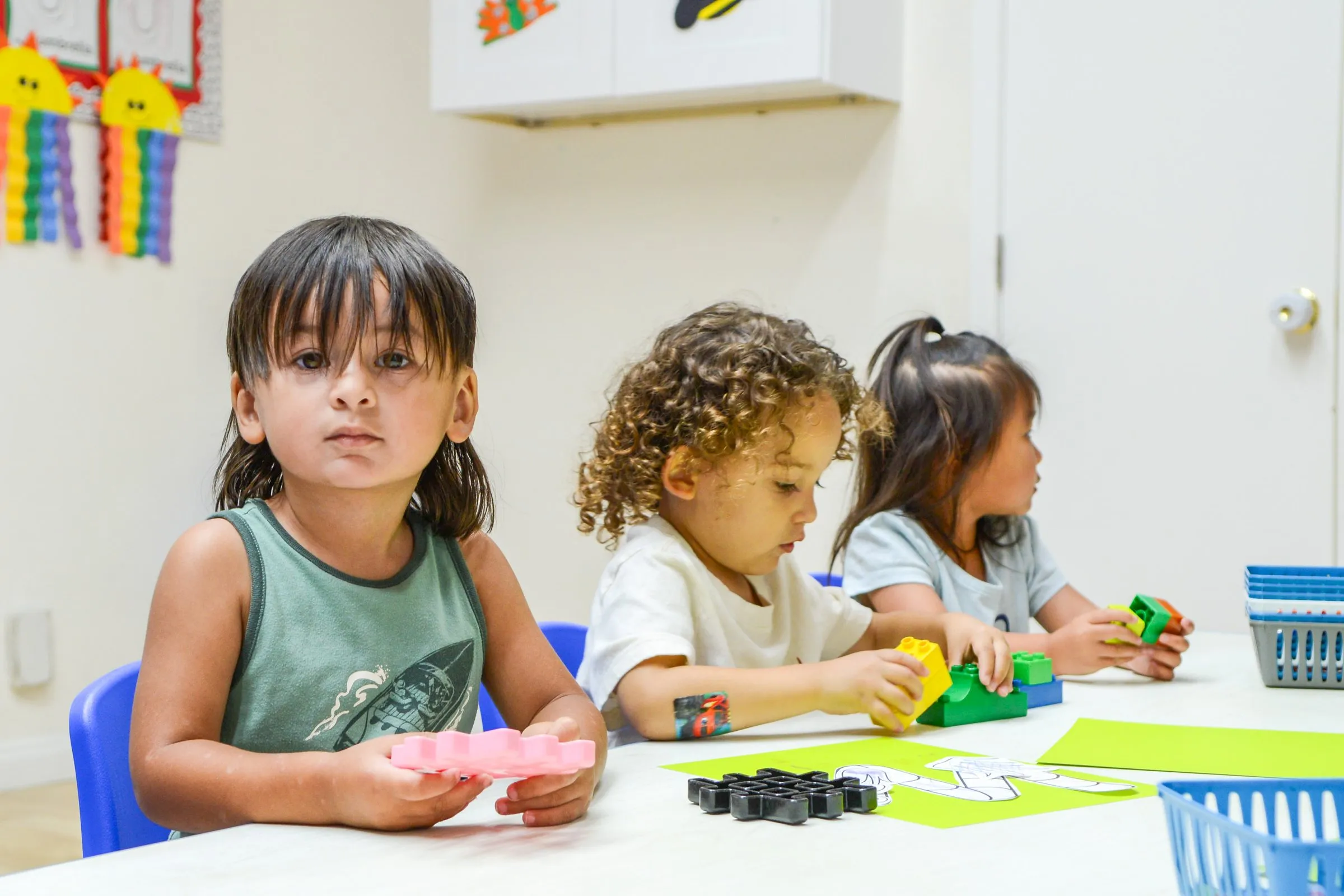 Three toddlers work with building toys at a table, one boy looking up at the camera.