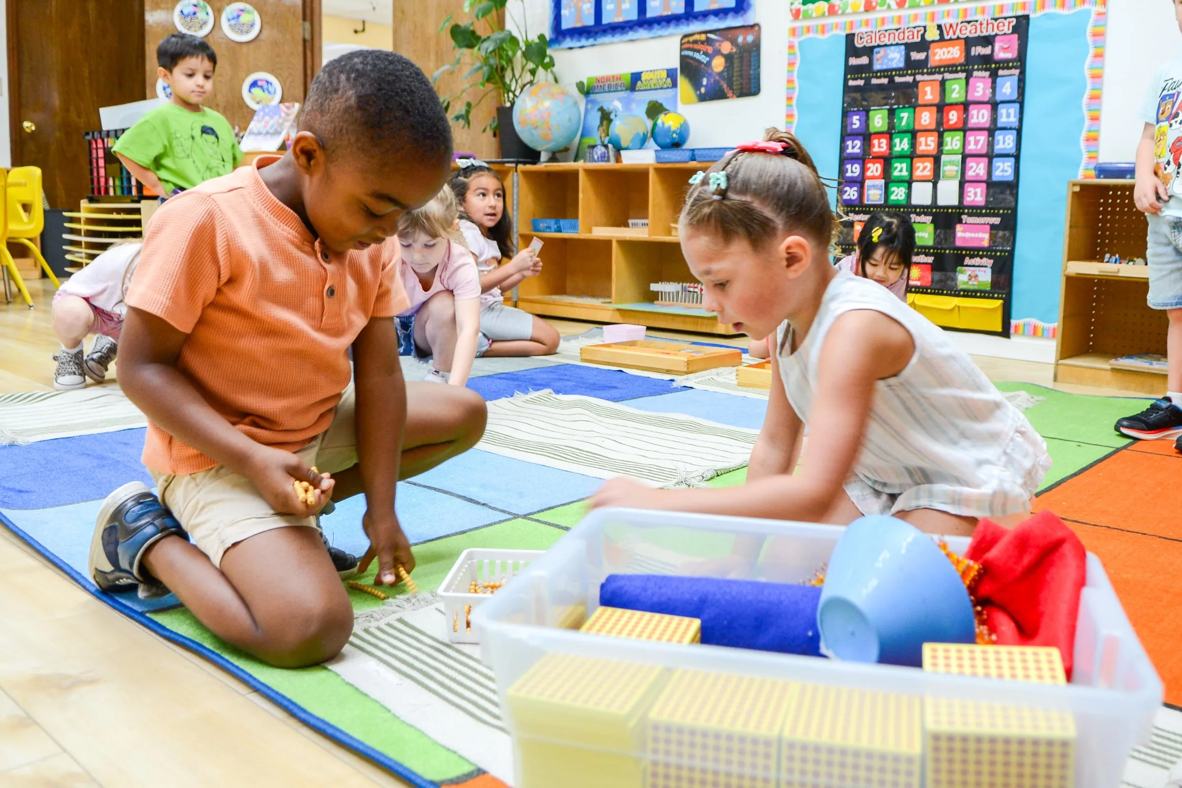 A boy and a girl work with Montessori bead materials on the classroom rug.