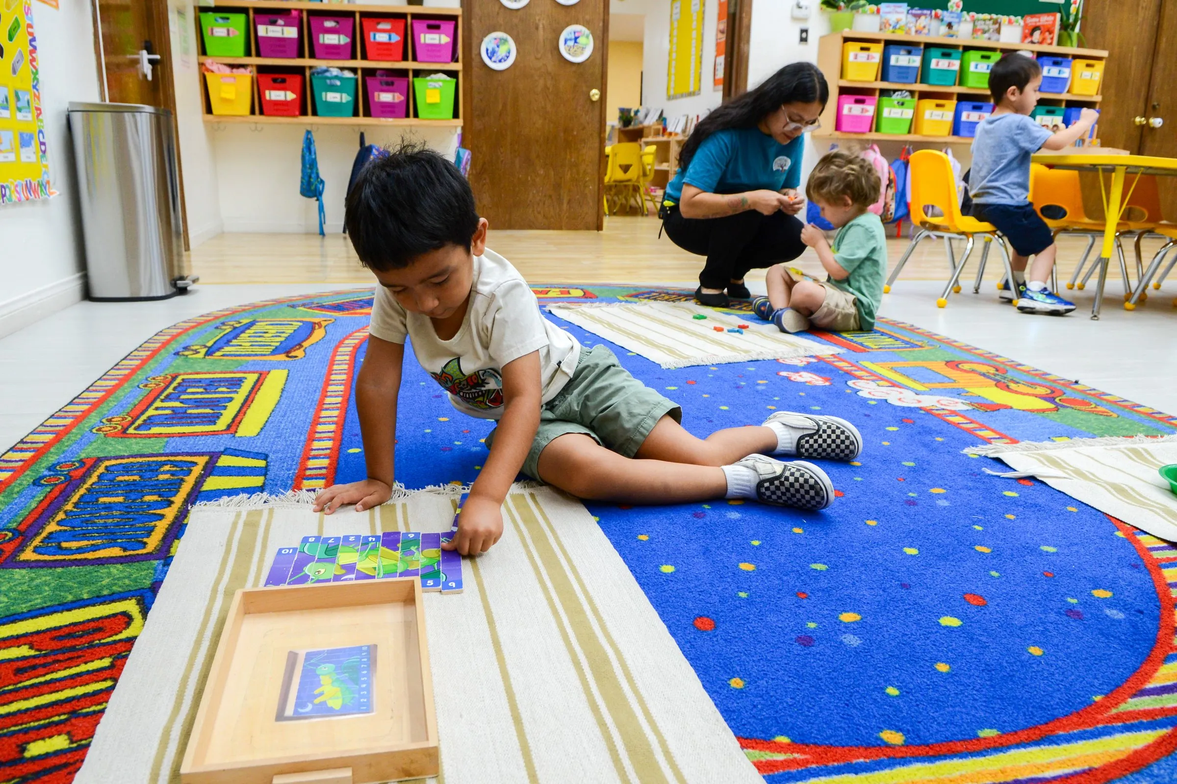 A boy kneels on a colorful rug working with materials from a wooden tray.