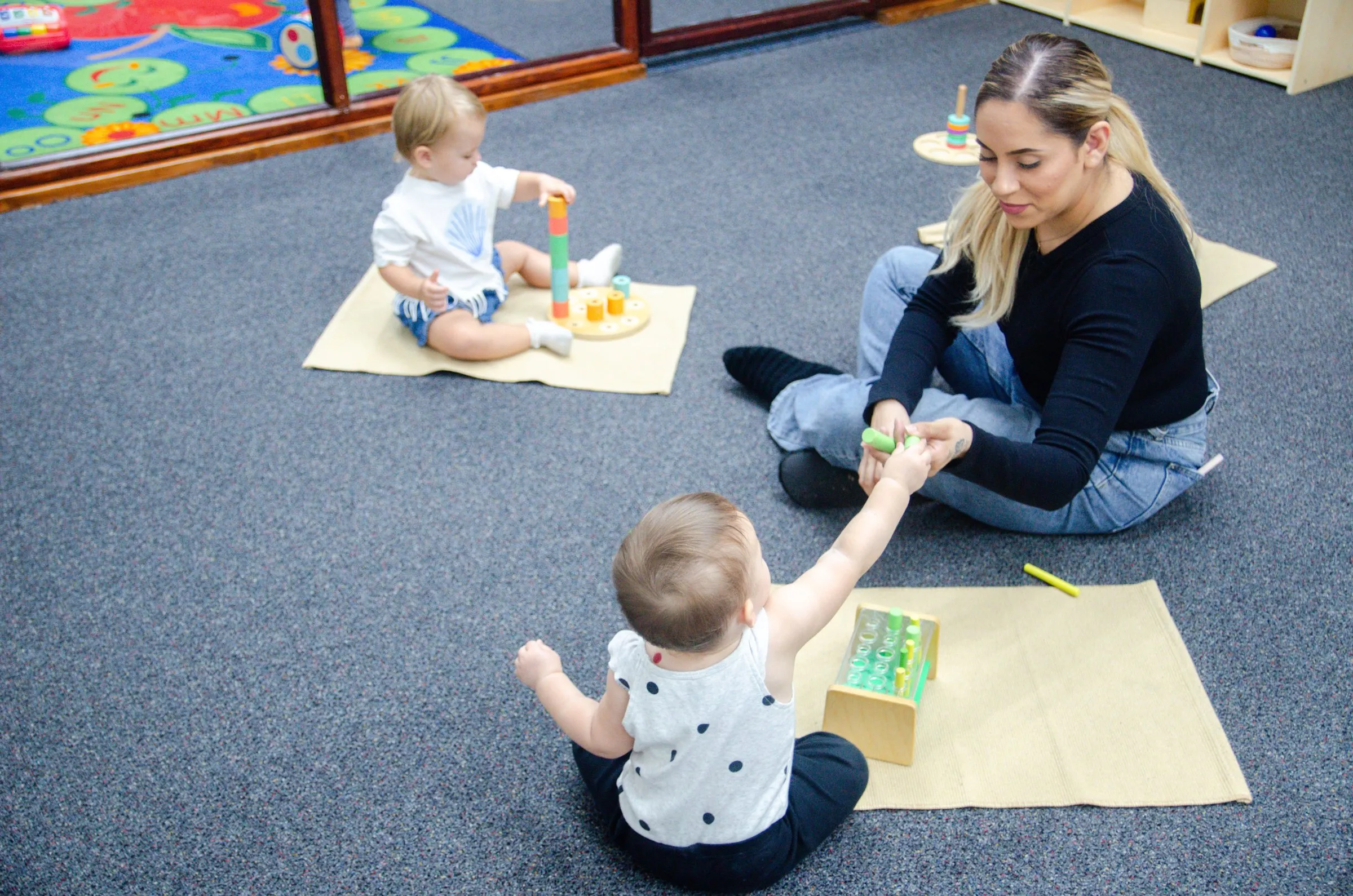 A teacher sits on the floor of the infant room with two babies, each playing on their own mat.