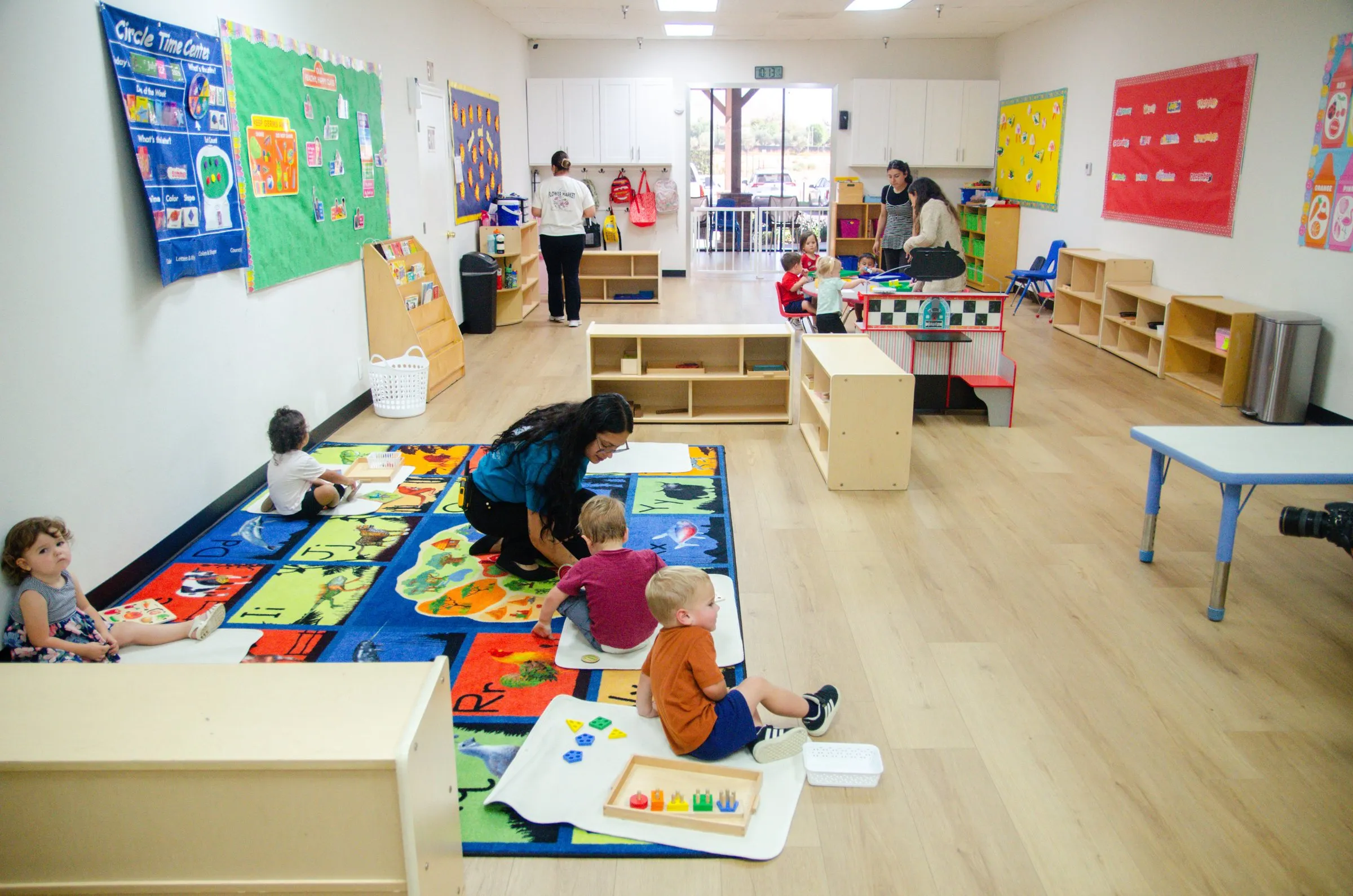 A bright infant and toddler room with a colorful rug, low shelves and a teacher playing with toddlers on the floor.
