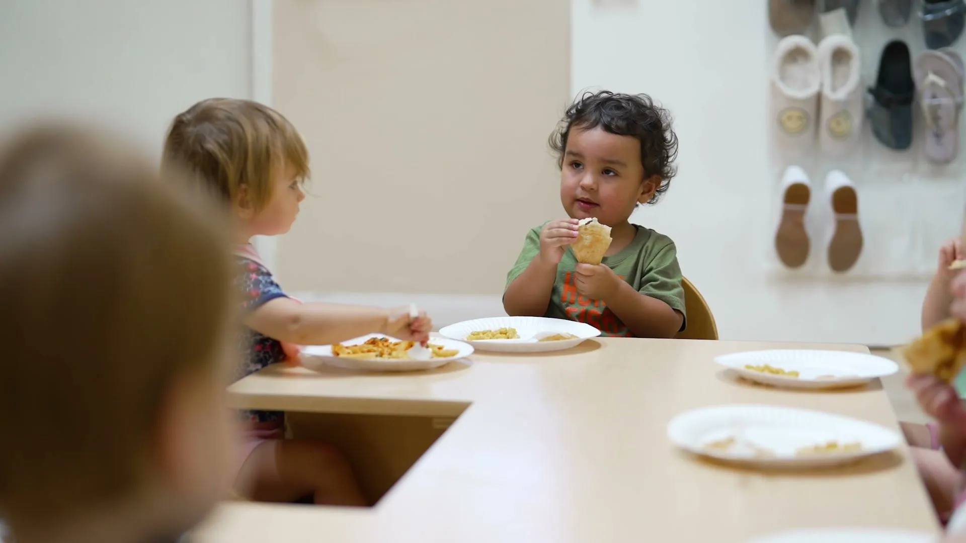 Toddlers eat lunch together at a low table, one smiling at the camera.