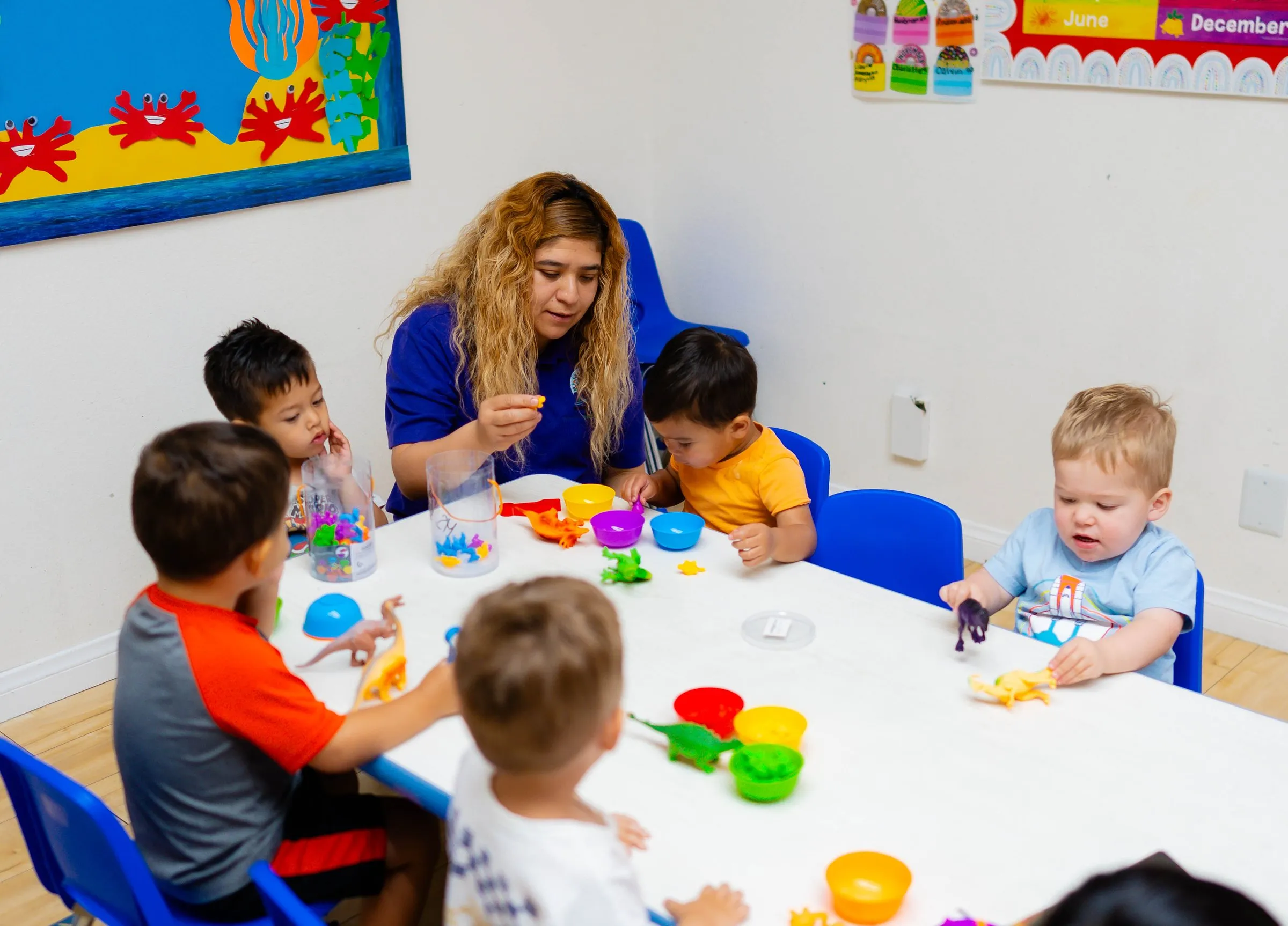 A teacher sits at a table with four toddlers working with colorful sorting cups.