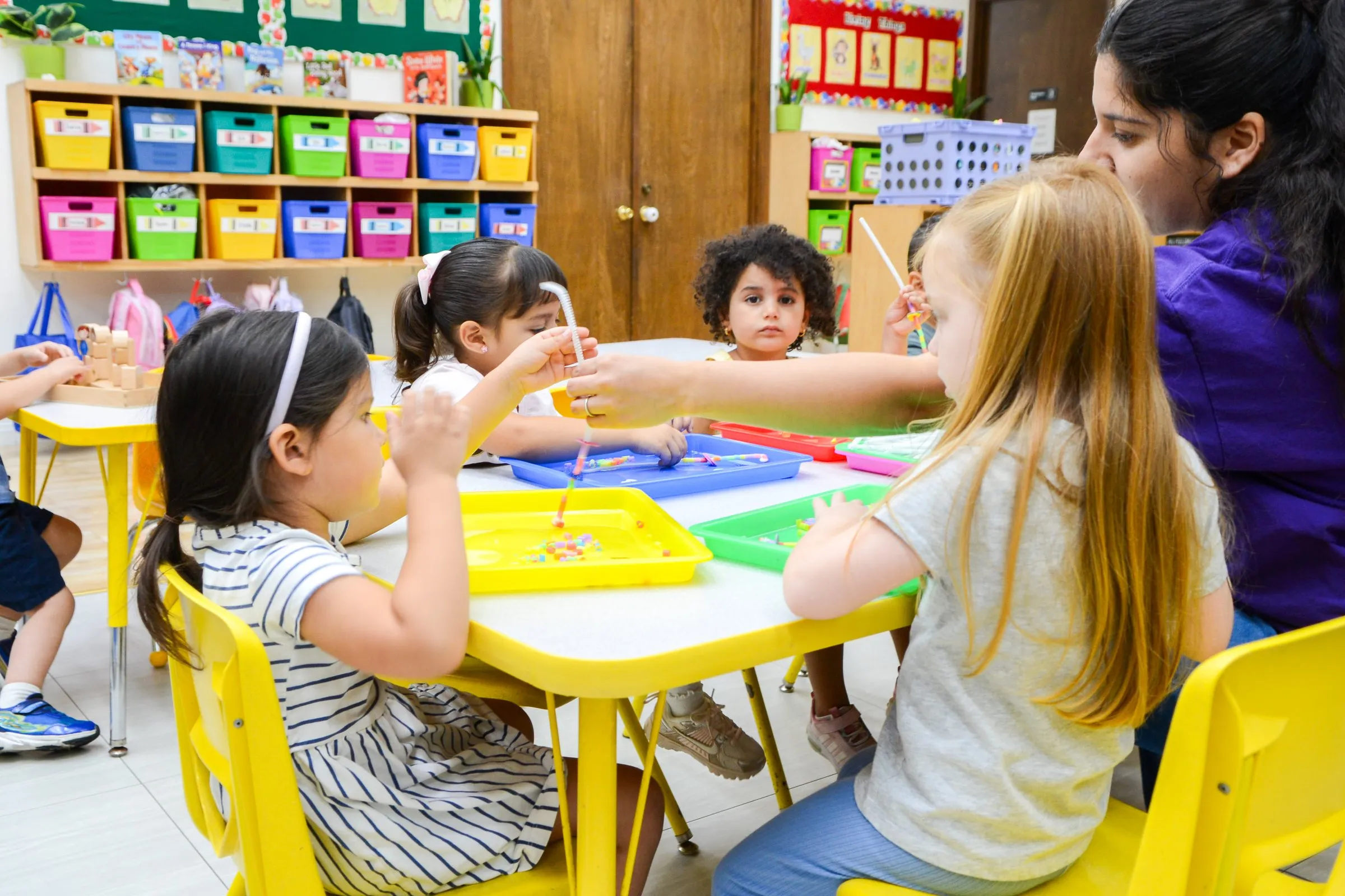 Children thread beads and sort small objects on colorful trays at a table with their teacher.