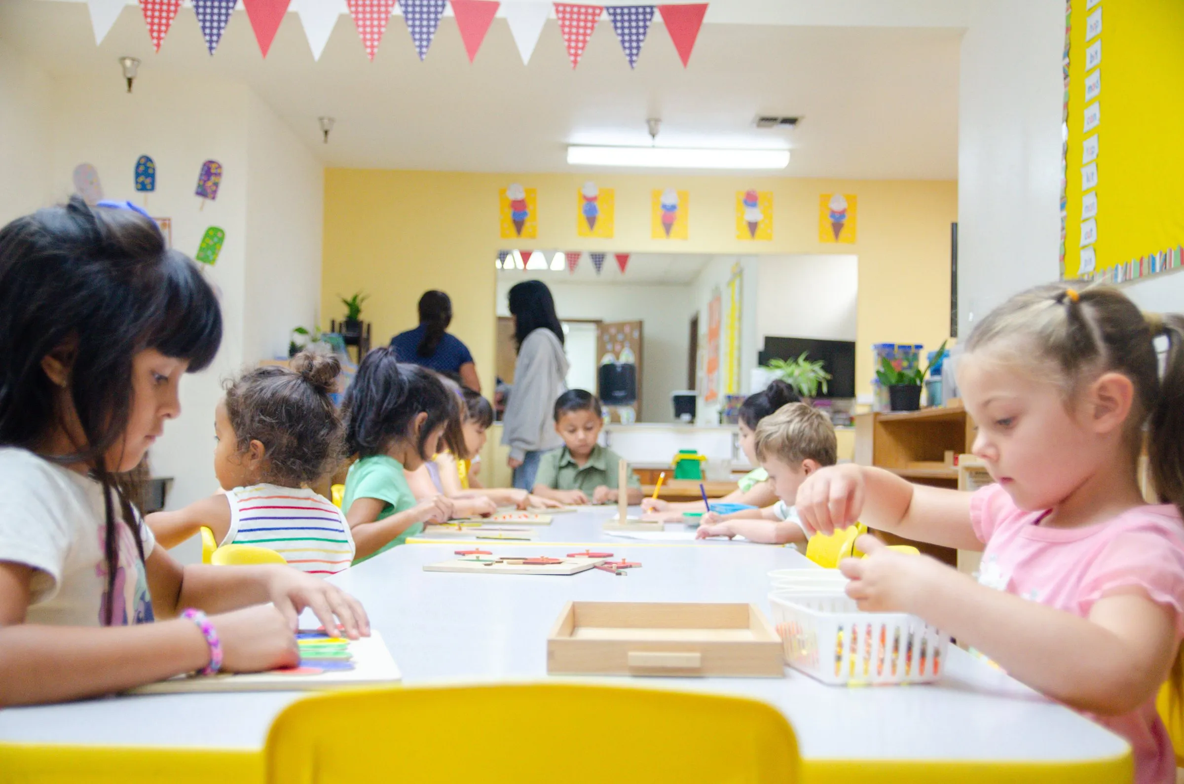 Children work on activities at a long table in a bright classroom decorated with bunting.